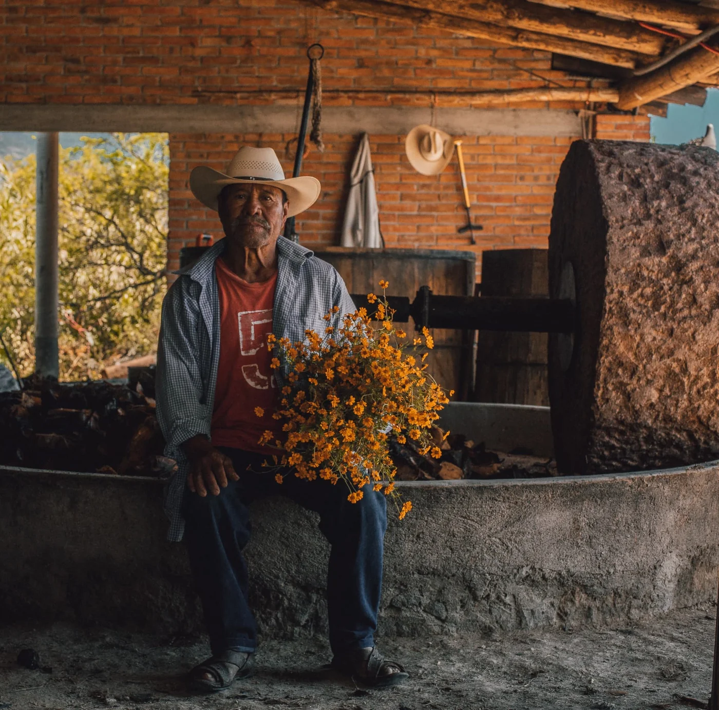 Maestro mezcalero sentado en el palenque con un ramo de flores de cempasúchil